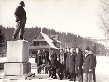A photograph of a statue in the town during winter. A group of men surround it, and a Zakopane style building is behind it.