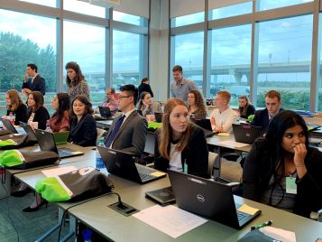 A group of students sitting in a room in front of their laptops. There are two large windows that meet in the corner behind them.