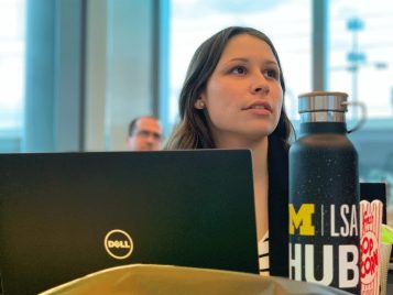 A young woman looks toward the front of the room. She has a Dell laptop open right in front of her, and she has a LSA Hub water bottle on her left.