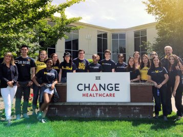 A group of students pose in a group around a sign marking Change Healthcare. They are all wearing U-M gear.