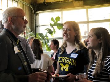 Two female students talk with an alumnus during a gathering. They are all wearing name tags and holding glasses and plates. There is a glass, roll-up garage door behind them, and a large green plant.