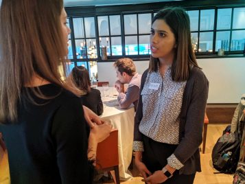 A female student wearing a name tag talks to an alumna in a large room with round tables covered with white table cloths. There are people sitting at the table in the background.