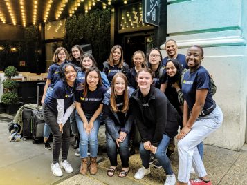 A cluster of 13 students posing in front of a hotel. The front row is crouching in front of the back row. They are wearing U-M gear.