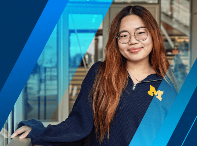 LSA Scholarship student Angelina Wong stands smiling at the entrance to the LSA Building atrium, smiling and wearing a navy blue long-sleeve quarter zip shirt with a maize block M on the chest. 