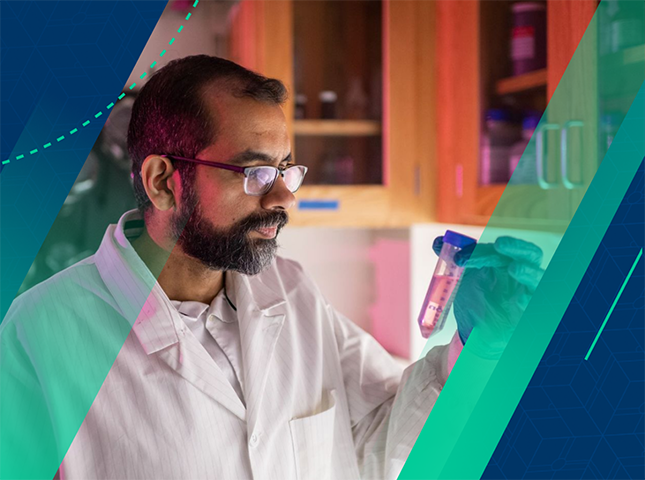 In purple lighting, Professor Omar Ahmed stands in front a lab counter with cabinets above, peering at a test tube filled with liquid. He is wearing eyeglasses, a lab coat and blue gloves. Other lab implements are attached to the rear wall of the room. 