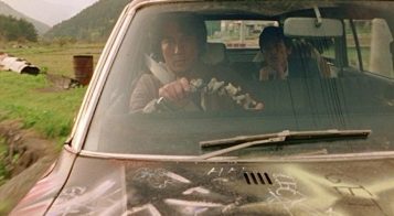 a stern looking man is driving through the countryside with a young man sitting in the backseat trying to talk to him