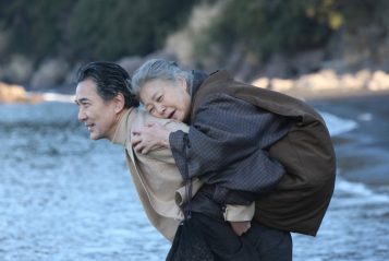 An elderly person is being carried piggyback by another person near a shoreline. Both appear content.