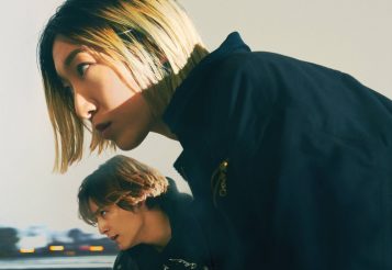 A man and a woman with short hair and wearing bacl jackets stare off into the distance