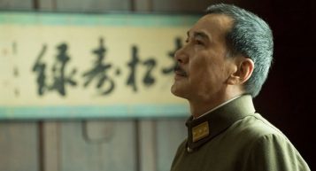 Man in military uniform looking upwards with a contemplative expression, standing in front of a wall with calligraphy.