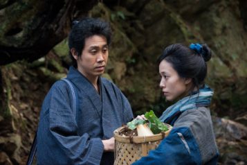 A woman carrying a basket of produce is staring off into the distance while a man stands in front of her and stares at her face. Both are wearing Japanese kimonos.