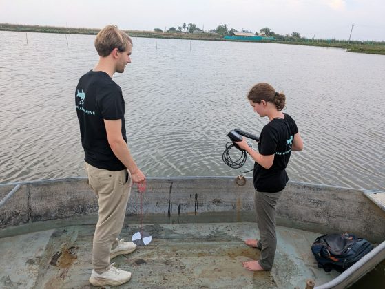 Annika and another individual, both wearing black shirts, are standing in a small aluminum boat on a body of water looking at a digital device