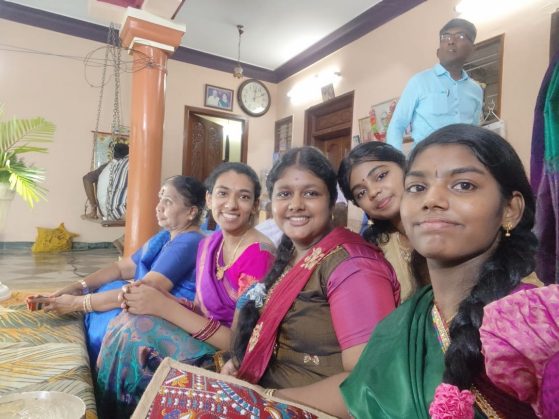 A group of people in colorful traditional attire sitting indoors, with a clock and plant visible in the background.