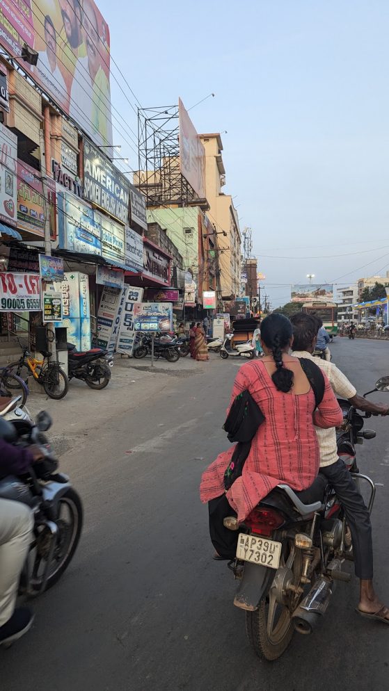 Two individuals ride on a motorcyle on a busy street in India 
