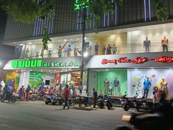 A bustling street scene in front of "Jaya Pa Textiles" with mannequins displayed above and people gathered below.