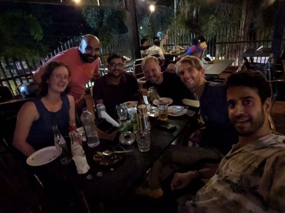 A group of six friends are sitting at a dinner table at night, smiling for a selfie