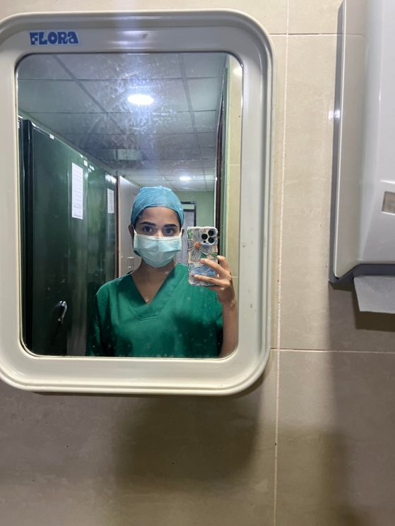 Varshini is taking a photo in the mirror wearing scrubs and a surgical face mask