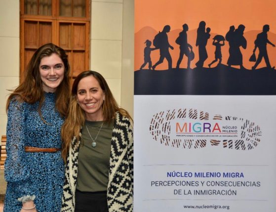 Two people stand by a banner reading "MIGRA NÚCLEO MILENIO: PERCEPCIONES Y CONSECUENCIAS DE LA INMIGRACIÓN"