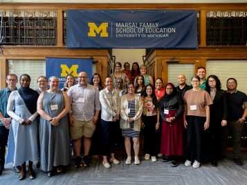 A group of approximately 25 people, a mix of men and women of various ages and ethnicities, pose for a group photo indoors. They are standing in two rows in front of a wooden staircase and a large banner that reads "Marsal Family School of Education, University of Michigan.