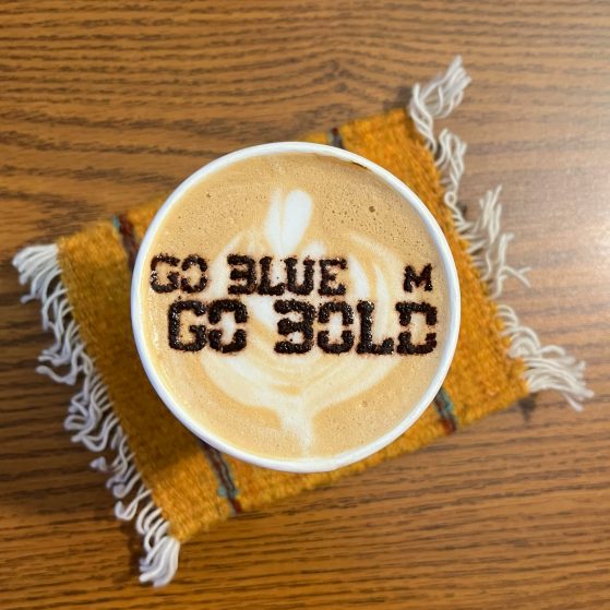 Image shows a cappuccino in a paper cup. There's cappuchino art on the top that spells out Go Blue Go Bold