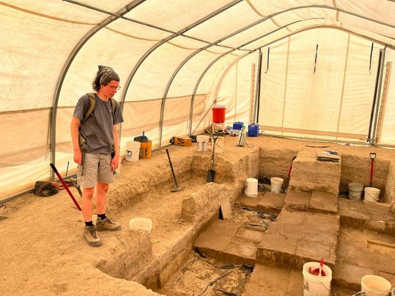 Caton inside a greenhouse-like structure covering a series of rectangular excavation holes surrounded by buckets, shovels, and other tools.