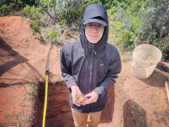 Caton is standing in an excavation hole that is about 3-feet deep and is marked off with yellow measuring tape with a whisk broom inside a bucket in the background.