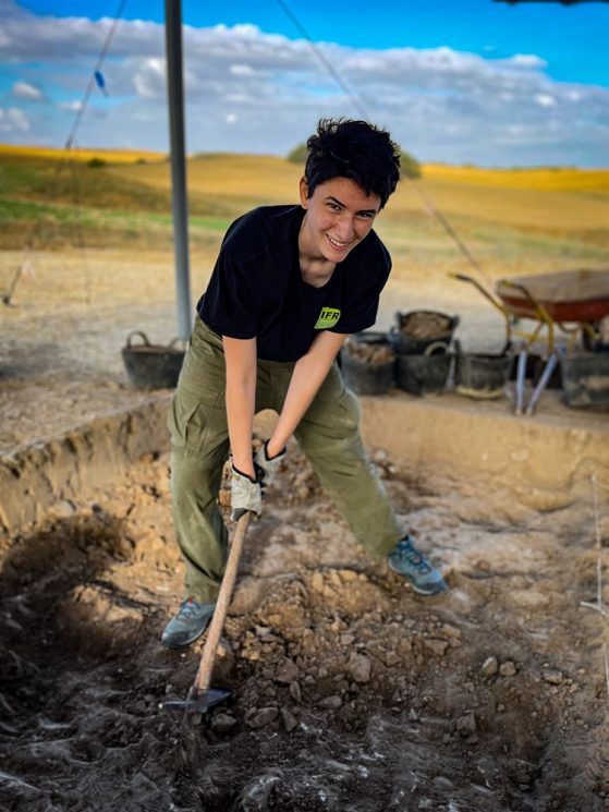 Welch excavating in a canopied, shallow site with a wheelbarrow and artifact-filled baskets on the rim.