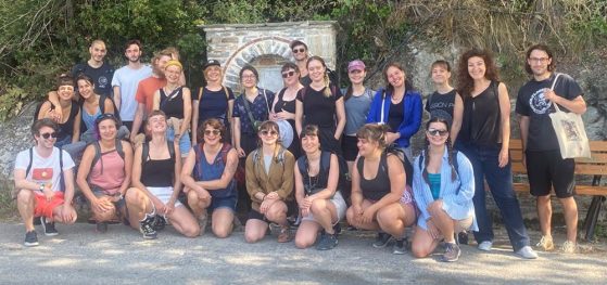 Group shot of 24 researchers standing in front of a forested hill containing a beige stone archway.