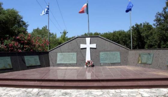 Monument with a raised floor and low stone walls containing plaques with a white cross with flowers at its base and three flags standing behind it.