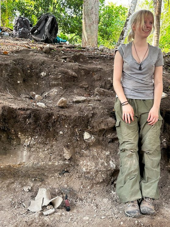 A smiling Humphrey standing in a deep excavation hole, leaning against its side, with an artifact and trowel at her feet and backpacks on the edge of the hole. 