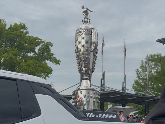 Large silver Borg-Warner Trophy displayed on a platform marked "109th RUNNING," with trees and flags in the background. 