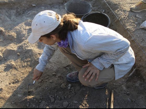 Pruette squatting down using a trowel to remove layers of soil at the site.