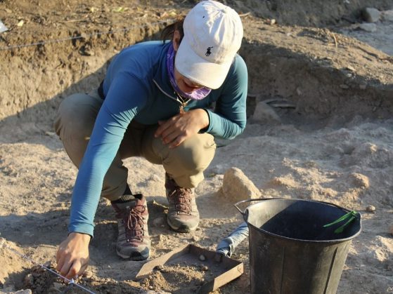 Pruette squatting down using a trowel and small sieve shovel to explore the site.