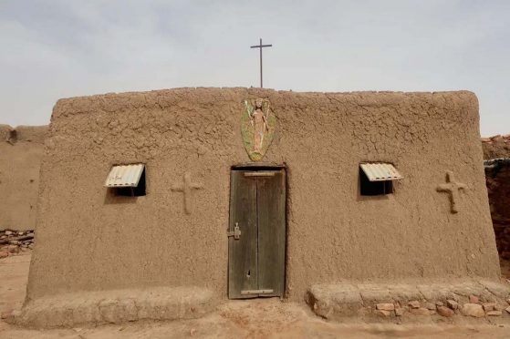 Simple stone church with wood door in center and a simple wooden cross on the roof. A single window and a stone cross appear on either side of the door. A statue on a green background is embedded in the wall above the door with the year 1986 underneath.