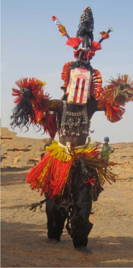 A Dogon adorned in ceremonial dress with a young boy in a baseball cap standing behind him watching.