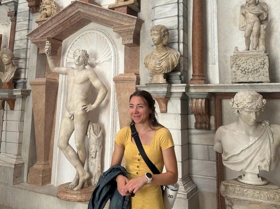 Person in a yellow dress stands in front of marble statues and busts in a grand hall with classical architectural details.