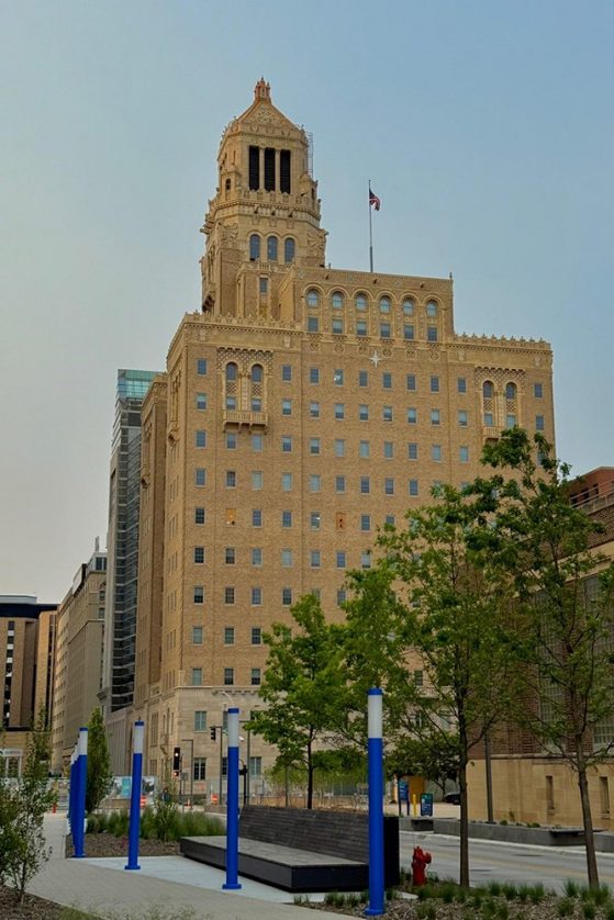 Tall, tan historic building with ornate tower, American flag, modern benches, blue poles, and green trees in foreground.