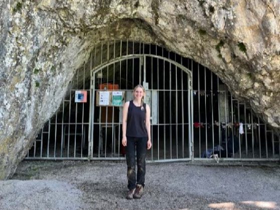 Heald, in her work clothes at the end of the day, standing in front of a stone opening filled with a silver barred fence that has double arched doors and signs hanging off it.