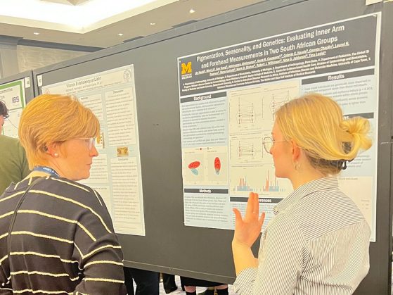 Two people discuss a University of Michigan research poster on pigmentation, seasonality, and genetics at a conference.