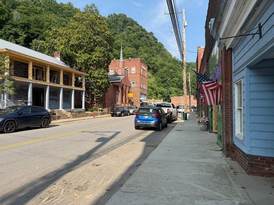 Small town street with parked cars, American flags, brick buildings, a house under construction, and green hills in the background.