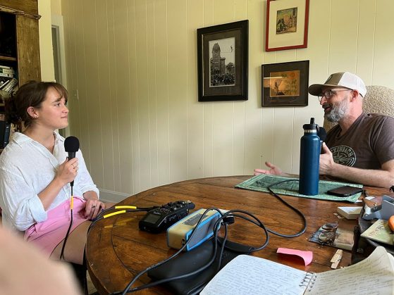 Two people sit at a wooden table with microphones and recording equipment, having a conversation in a cozy, decorated room.