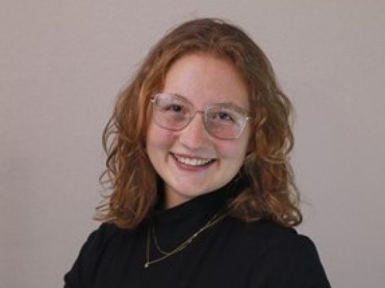 A headshot of the red-headed, smiling Tutak wearing a black sweater with a double necklace and her glasses.