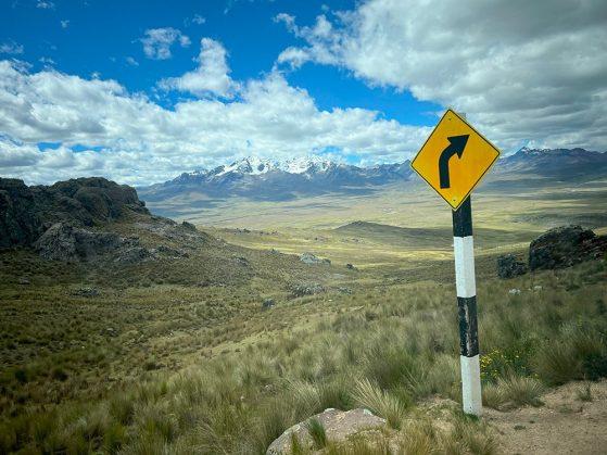 Yellow right-turn road sign on a post stands in grassy highlands, with mountains and cloudy skies in the background.