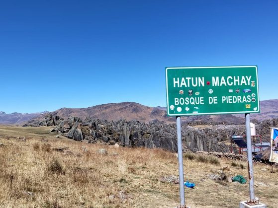 Green road sign reading "Hatun Machay Bosque de Piedras" near rocky formations under a clear blue sky in a dry landscape.