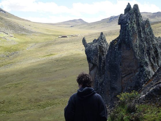Person with curly hair in a black jacket stands near jagged rocks, overlooking a vast, grassy valley with distant buildings.