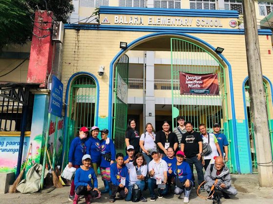 A group of people pose in front of the Balara Elementary School gate with a Dengue Awareness Month banner visible above them.