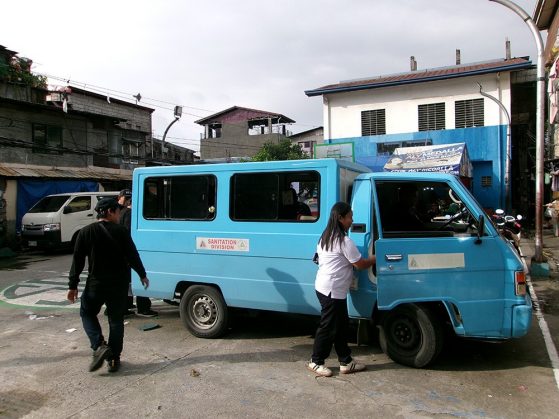 A blue sanitation division truck is parked. A woman opens the passenger door while a man walks nearby in an urban area.