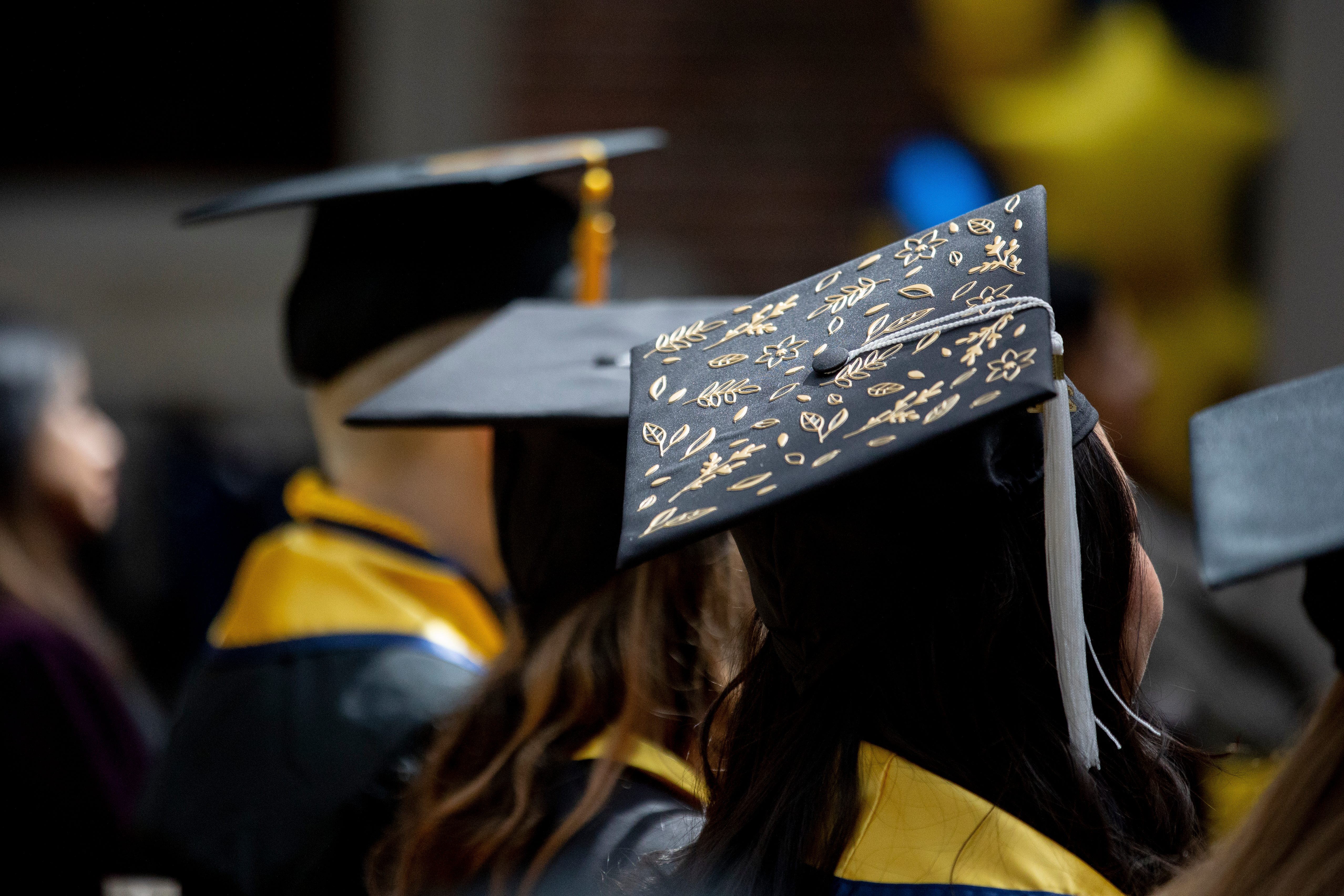 Large group of people, some in graduation gowns and caps, in a large room facing a person speaking at a podium