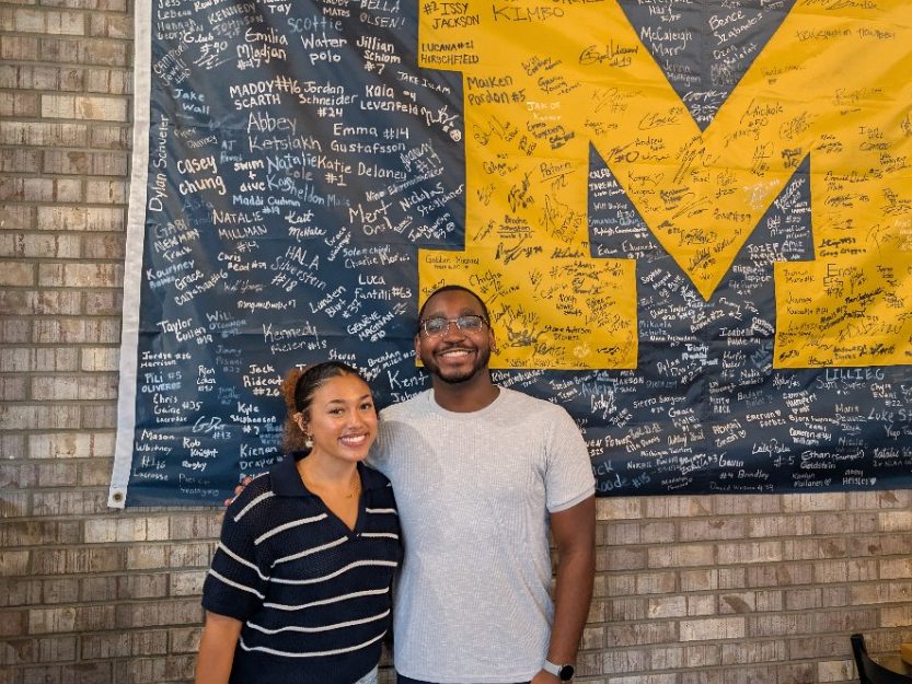 BLI intern standing with their supervisor in front of a large maize and blue Michigan M flag covered in signatures. 