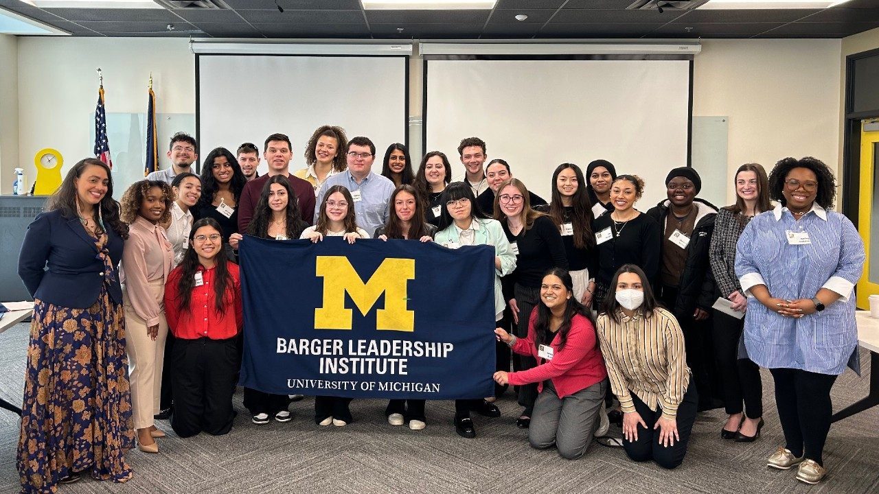group of two dozen students and four speakers standing in a group holding a blue Michigan Barger Leadership Institute flag.
