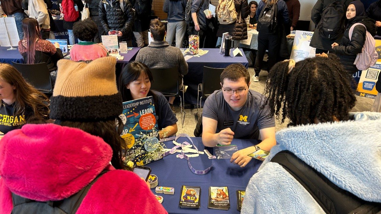Two BLI students sitting at a table representing the Barger Leadership Institute. There are two students with their backs to the camera talking to the students at the table. The rest of the room is full of people at other tables. 